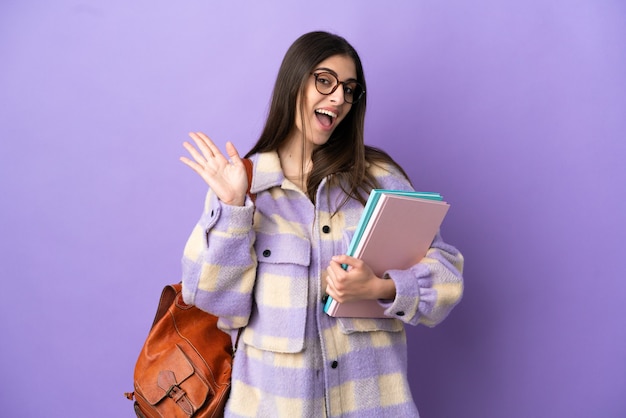 Mujer joven estudiante aislada sobre fondo púrpura saludando con la mano con expresión feliz