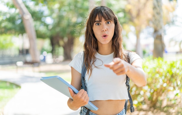 Mujer joven estudiante al aire libre sorprendida y apuntando al frente