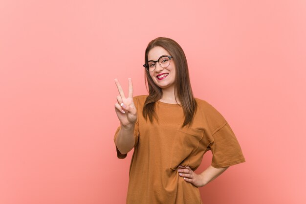 Mujer joven estudiante con anteojos mostrando signo de victoria y sonriendo ampliamente.