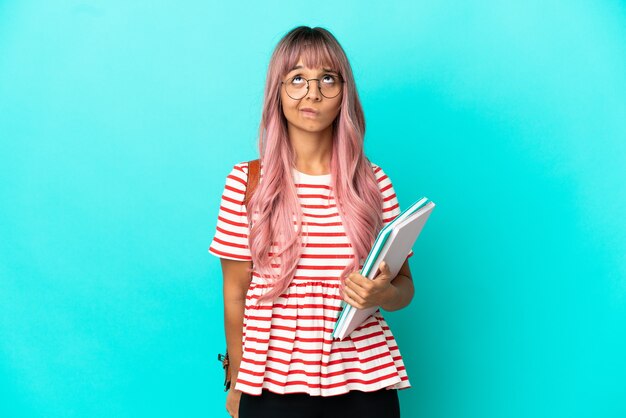 Mujer joven estudiante con cabello rosado aislado sobre fondo azul y mirando hacia arriba