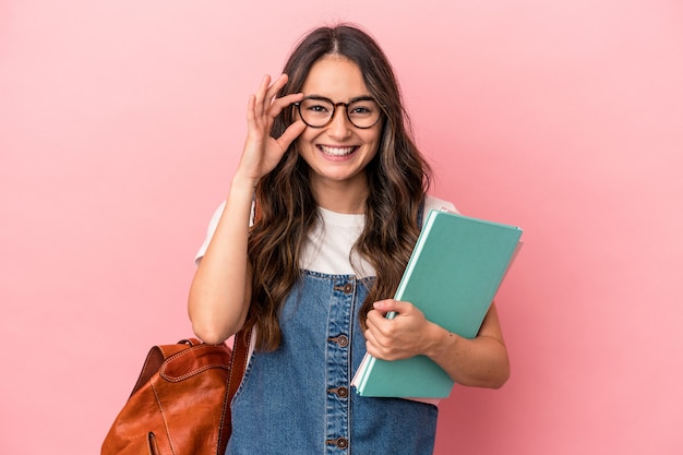 Mujer joven estudiante caucásica aislada sobre fondo rosa emocionado manteniendo el gesto ok en el ojo.