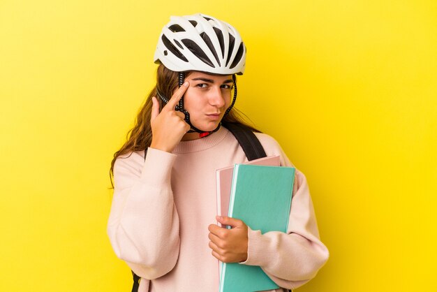 Mujer joven estudiante caucásica con un casco de bicicleta aislado sobre fondo amarillo apuntando al templo con el dedo, pensando, centrado en una tarea.