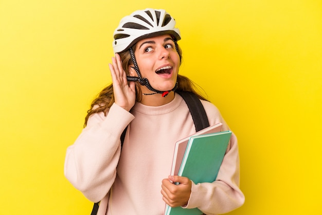 Mujer joven estudiante caucásica con un casco de bicicleta aislado sobre fondo amarillo tratando de escuchar un chisme.