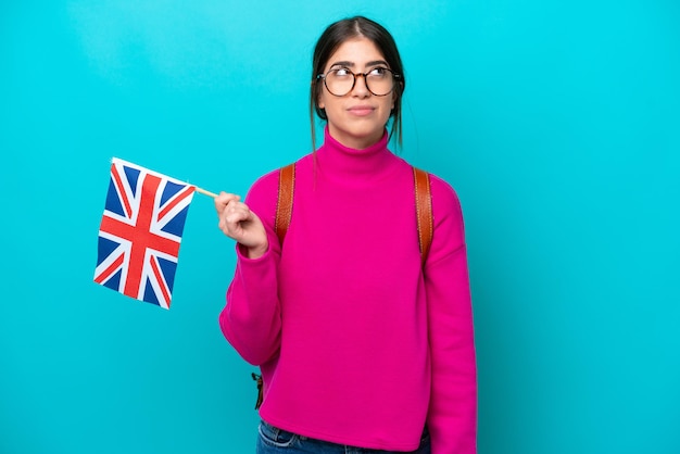 Mujer joven estudiante caucásica sosteniendo la bandera inglesa aislada de fondo azul y mirando hacia arriba