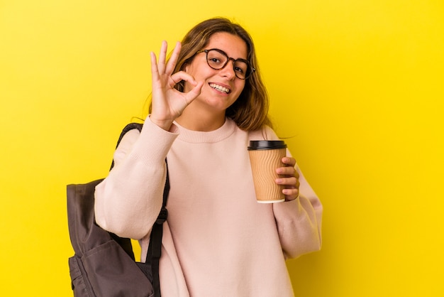 Mujer joven estudiante caucásica sosteniendo café aislado sobre fondo amarillo alegre y confiado mostrando gesto ok.