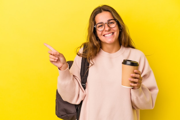 Mujer joven estudiante caucásica sosteniendo café aislado sobre fondo amarillo sonriendo y apuntando a un lado, mostrando algo en el espacio en blanco.