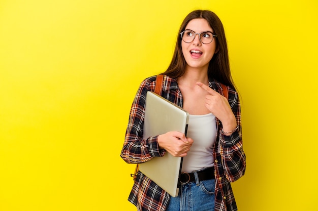 Mujer joven estudiante caucásica sosteniendo una computadora portátil aislada en puntos de pared rosa con el dedo pulgar lejos, riendo y despreocupado