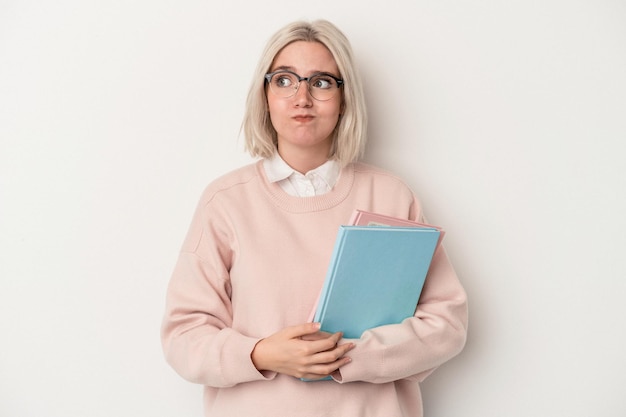 Mujer joven estudiante caucásica sosteniendo libros aislados sobre fondo blanco confundida, se siente dudosa e insegura.