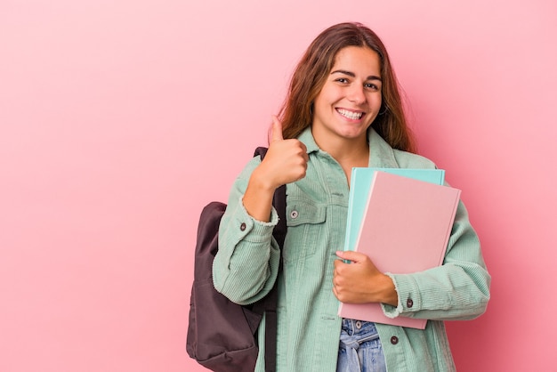 Mujer joven estudiante caucásica sosteniendo libros aislados sobre fondo rosa sonriendo y levantando el pulgar hacia arriba
