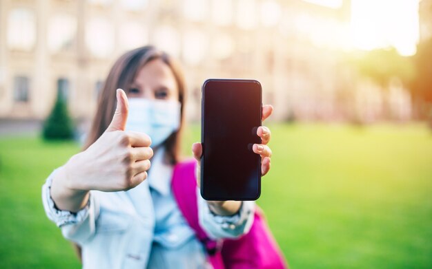 Mujer joven estudiante confiada en una máscara médica está apuntando en la pantalla del teléfono inteligente en blanco y de pie en el campus universitario