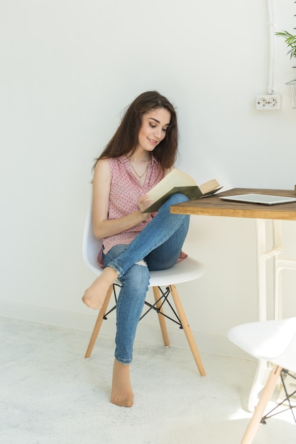Mujer joven estudiante está leyendo un libro, mientras está sentada en una silla blanca en una habitación blanca