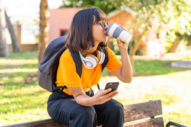 Mujer joven estudiante gana un parque con teléfono móvil y sosteniendo un café para llevar