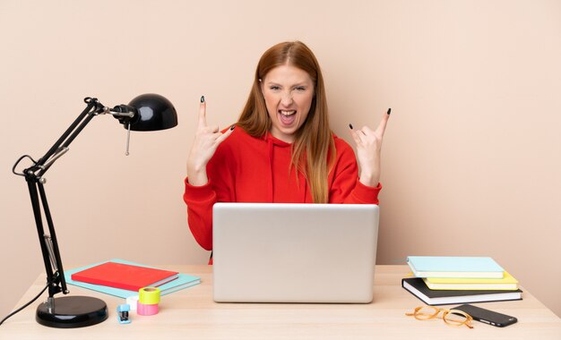 Mujer joven estudiante en un lugar de trabajo con una computadora portátil haciendo gesto de rock