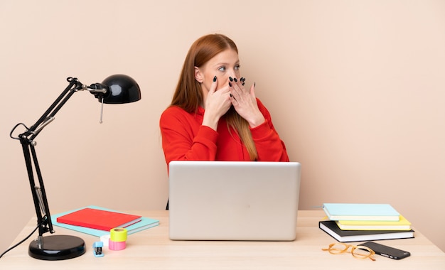 Mujer joven estudiante en un lugar de trabajo con un portátil cubriendo la boca y mirando hacia un lado