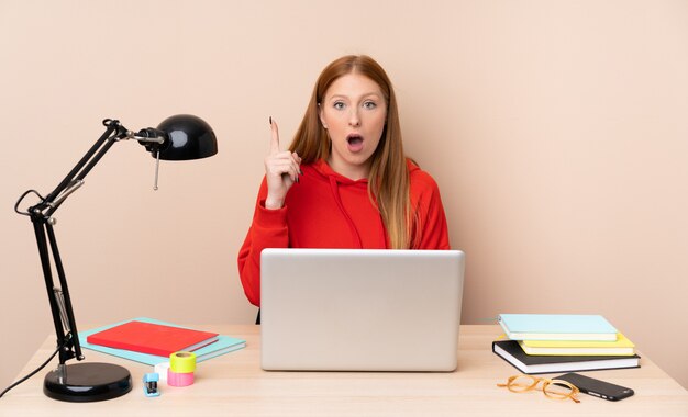 Mujer joven estudiante en un lugar de trabajo con un portátil pensando en una idea apuntando el dedo hacia arriba