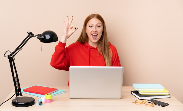 Mujer joven estudiante en un lugar de trabajo con un portátil sorprendido y mostrando signo ok
