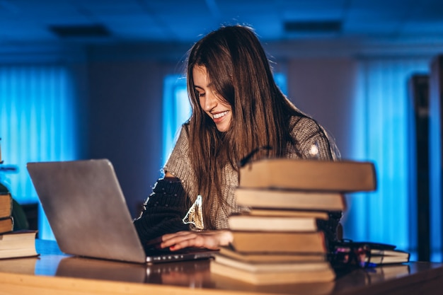 Mujer joven estudiante en la noche se sienta en una mesa en la biblioteca con una pila de libros y trabaja en una computadora portátil. Preparándose para el examen