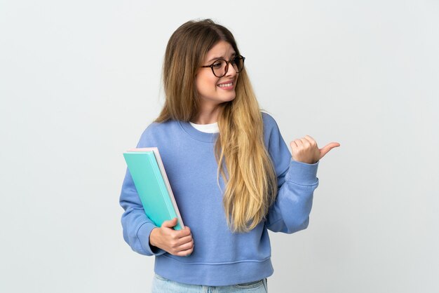 Mujer joven estudiante rubia aislada en la pared blanca apuntando hacia el lado para presentar un producto