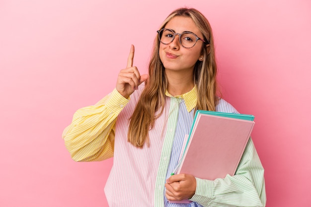 Mujer joven estudiante rubia caucásica sosteniendo libros aislados mostrando el número uno con el dedo.