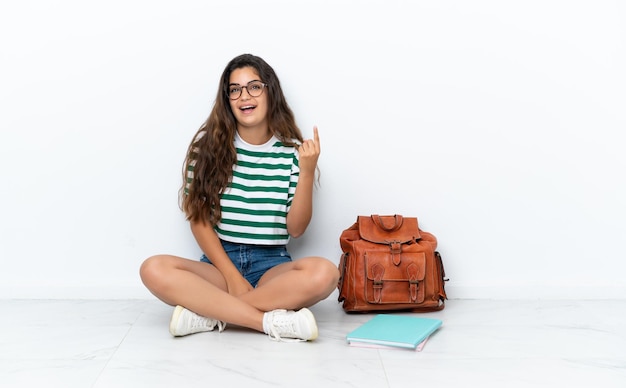 Mujer joven estudiante sentada en el suelo aislada de fondo blanco apuntando hacia arriba y sorprendida