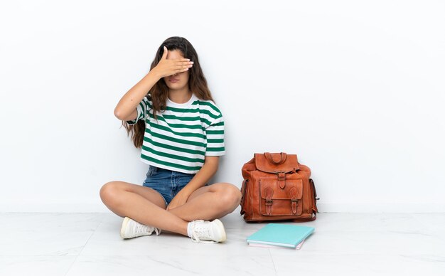 Mujer joven estudiante sentada uno en el suelo aislado sobre fondo blanco cubriendo los ojos con las manos. No quiero ver algo