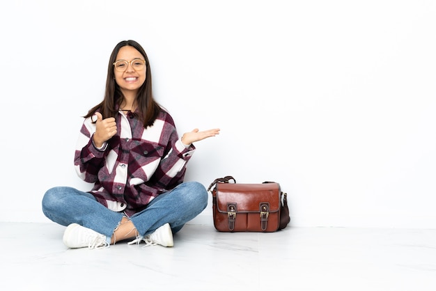 Mujer joven estudiante sentada en el suelo sosteniendo copyspace imaginario en la palma para insertar un anuncio y con los pulgares hacia arriba