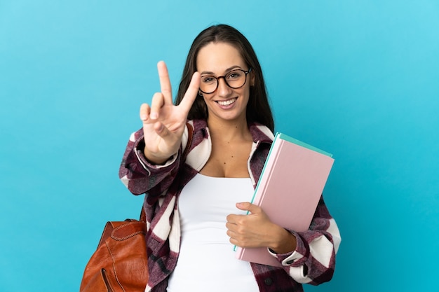 Mujer joven estudiante sobre pared aislada sonriendo y mostrando el signo de la victoria