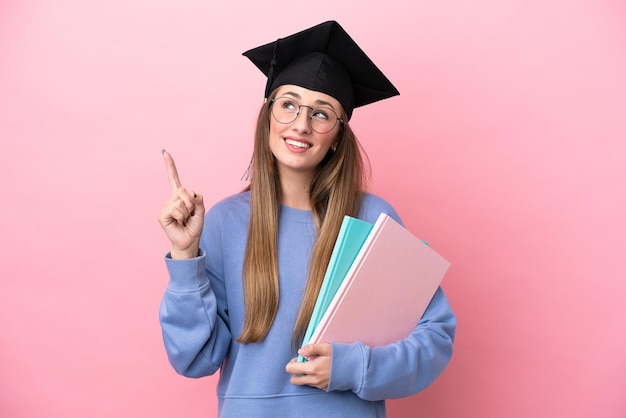 Mujer joven estudiante con un sombrero de posgrado aislado en un fondo rosado que señala una gran idea