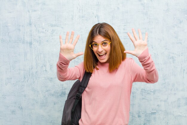 Mujer joven estudiante sonriendo y mirando amigable, mostrando el número diez o décimo con la mano hacia adelante, contando contra la pared de la pared del grunge