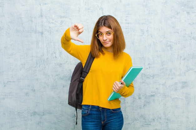 Mujer joven estudiante triste, decepcionada o enojada, mostrando los pulgares hacia abajo en desacuerdo, sintiéndose frustrada contra el fondo de la pared del grunge