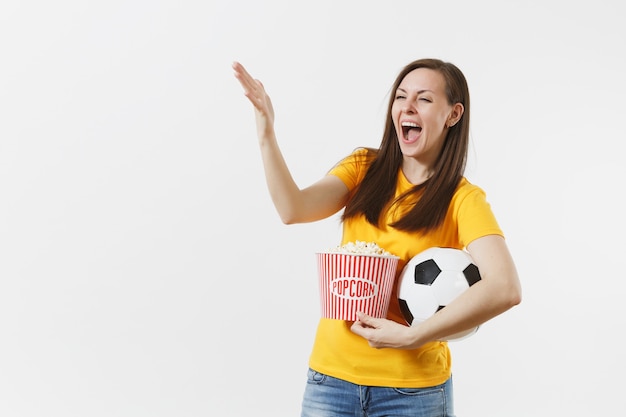 Mujer joven europea llena de alegría, aficionado al fútbol o jugador en uniforme amarillo con balón de fútbol, cubo de palomitas de maíz aislado sobre fondo blanco. Deporte, jugar al fútbol, animar, concepto de estilo de vida de la gente de los fanáticos
