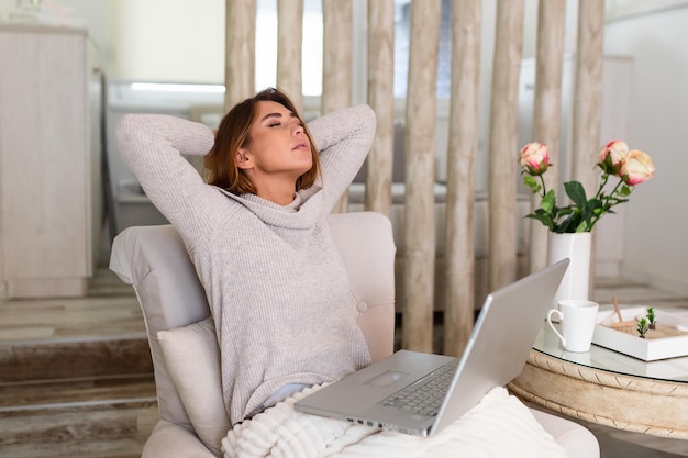 Mujer joven con exceso de trabajo sentada en su casa con una computadora portátil frente a ella estirando los brazos por encima de la cabeza con los ojos cerrados para relajar los músculos, reducir la tensión, mejorar la visión,