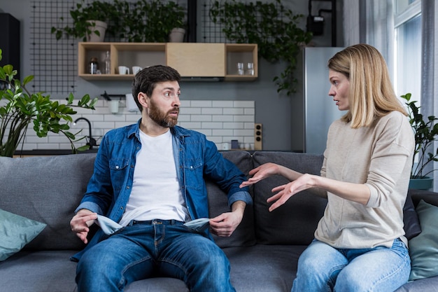 Una mujer joven le exige dinero a su esposo, el hombre no muestra los bolsillos vacíos En casa sentado en el sofá Peleas conflictos malentendidos Crisis de dinero