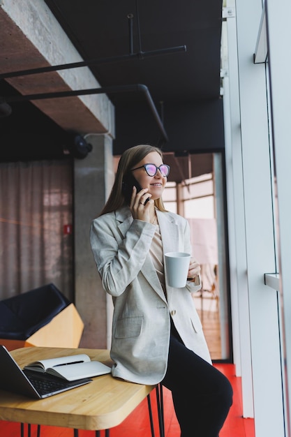 Mujer joven exitosa resolviendo problemas de negocios durante una conversación telefónica sentada en un café moderno y trabajando con una computadora portátil Lindo interior está disfrutando de su tiempo libre en el café