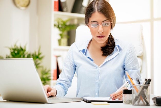 Mujer joven exitosa trabajando en su escritorio en la oficina. Ella está usando una computadora portátil y analizando el informe comercial.