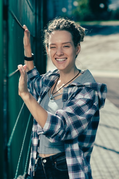 Mujer joven expresiva sonriendo felizmente y guiñando un ojo mientras está de pie cerca de la valla de tela metálica del campo de deportes