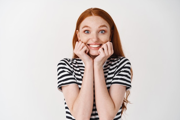 Mujer joven extremadamente feliz y sorprendida con el pelo rojo, tocando la cara y sonriendo asombrado, recibiendo un regalo sorpresa, pared blanca