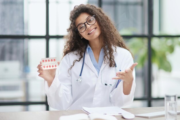 Mujer joven farmacéutica sosteniendo pastillas mientras está sentada en la mesa en la oficina