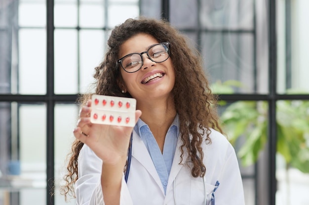 Mujer joven farmacéutica sosteniendo pastillas mientras está sentada en la mesa en la oficina