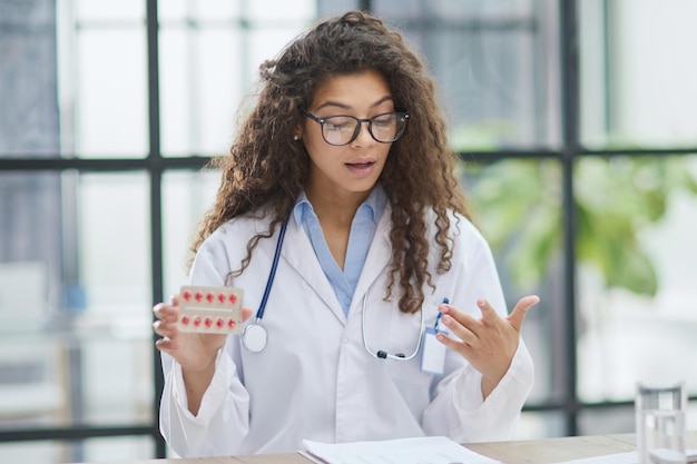 Mujer joven farmacéutica sosteniendo pastillas mientras está sentada en la mesa en la oficina