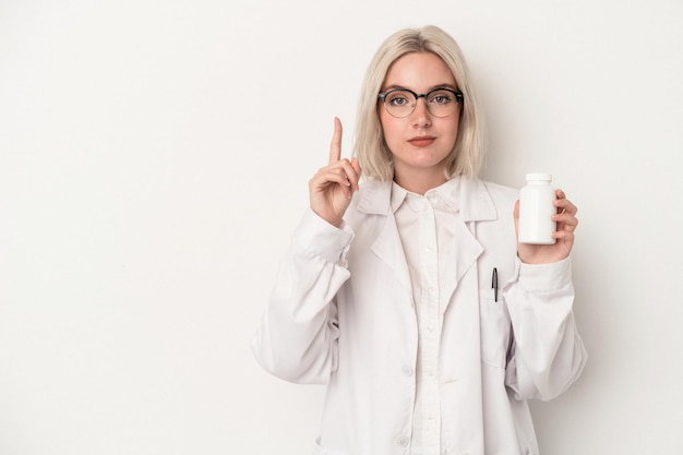 Mujer joven farmacéutica sosteniendo píldoras aisladas sobre fondo blanco mostrando el número uno con el dedo