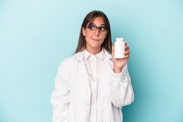 Mujer joven farmacéutico con píldoras aisladas sobre fondo azul confundido, se siente dudoso e inseguro.