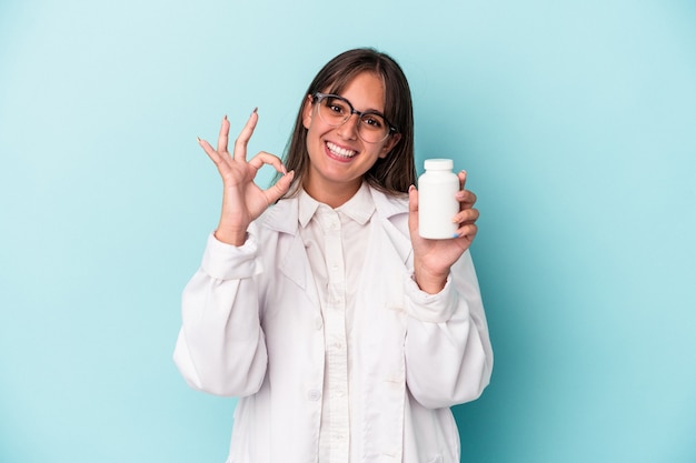 Mujer joven farmacéutico sosteniendo píldoras aisladas sobre fondo azul alegre y confiado mostrando gesto ok.