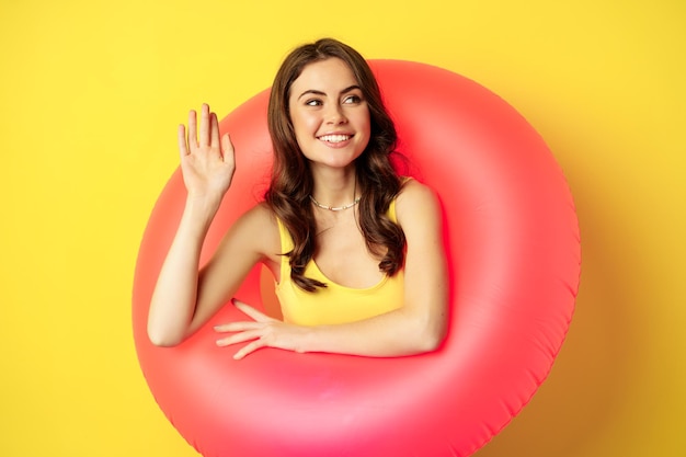 Mujer joven feliz con anillo de natación rosa saludando con la mano y diciendo hola disfrutando de las vacaciones de verano en la playa...