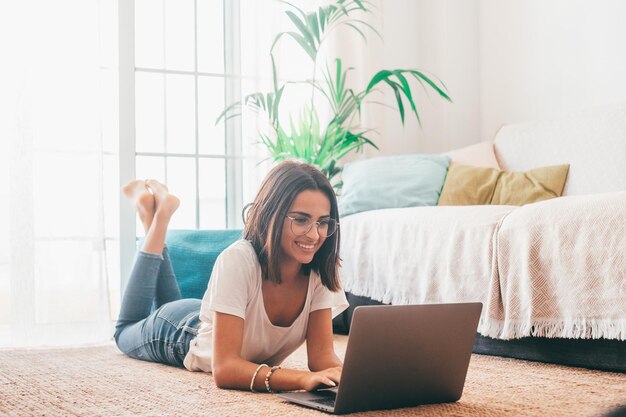 Foto mujer joven feliz en anteojos escribiendo usando la computadora portátil mientras está acostada sobre la alfombra en la sala de su casa. bella dama pasar tiempo libre usando la computadora portátil. freelancer trabajando desde casa