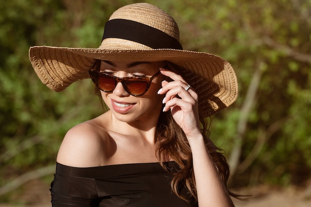 Mujer joven feliz de apariencia caucásica en un primer plano de sombrero de paja en gafas de sol posando en un día soleado de verano en la playa