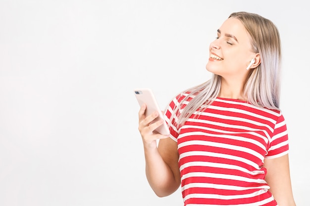 Mujer joven feliz en auriculares está escuchando música con teléfonos inteligentes, bailando, divirtiéndose y sonriendo. Fondo blanco, aislado.