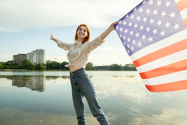 Mujer joven feliz con la bandera nacional de Estados Unidos sobre sus hombros con altos edificios de la ciudad en el fondo celebrando el día de la independencia de Estados Unidos