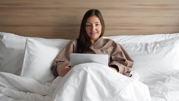 Mujer joven feliz en bata de baño con tableta acostado en la cama de matrimonio en la vista frontal del dormitorio moderno