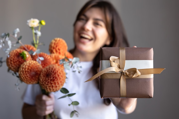 Mujer joven feliz con una caja de regalo y un ramo de flores de crisantemo sobre un fondo gris.